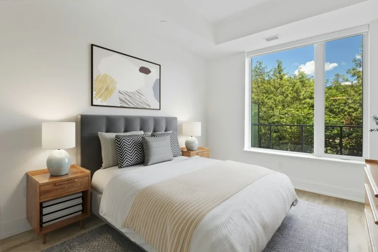 Bedroom with a grey headboard, white bedding, two lamps and a large window onto trees