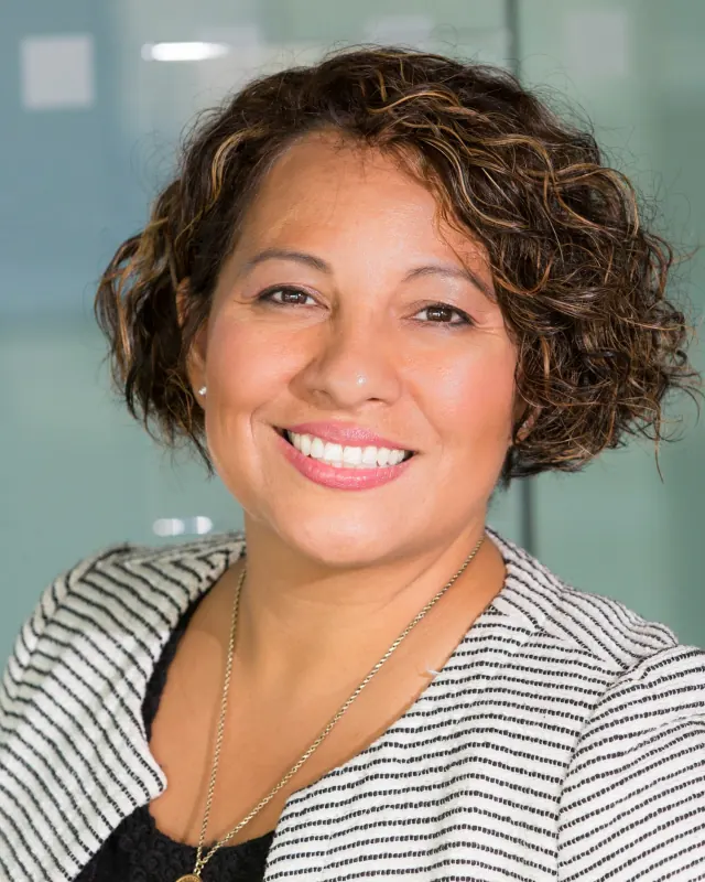 Danielle, a smiling woman with short curly hair in a striped jacket, photographed indoors against a glass wall