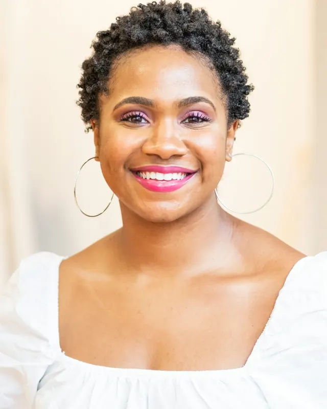 Imani, a woman with short natural hair and hoop earrings in a white blouse, smiling at the camera