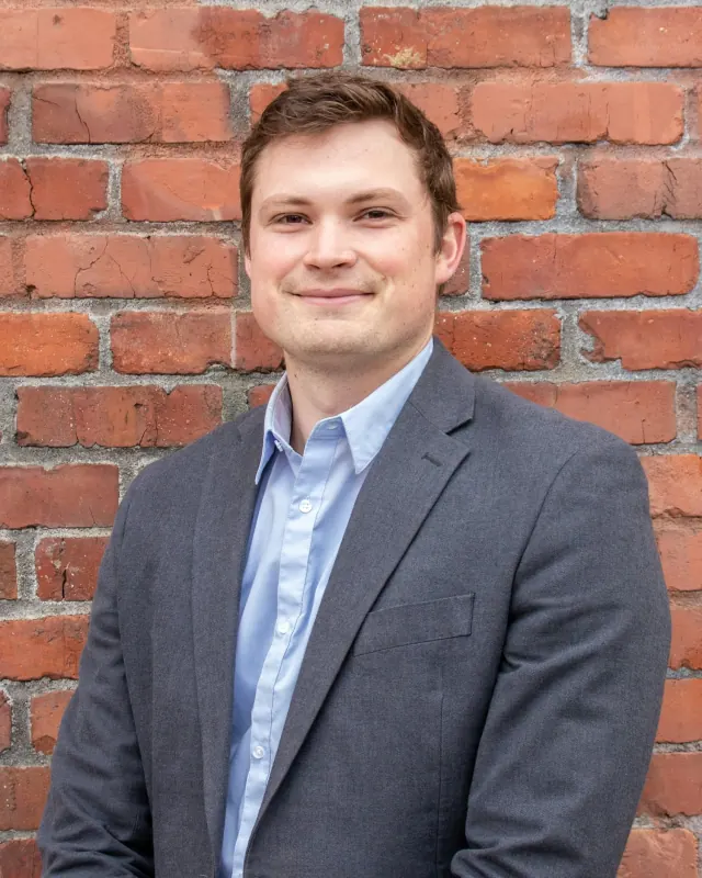Owen, a man in his thirties in a navy blazer and light blue shirt, smiling slightly in front of a brick wall