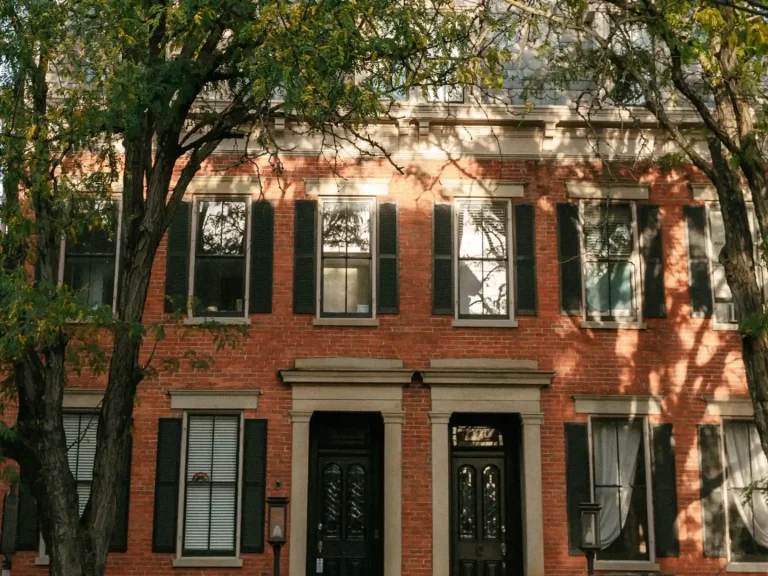 Two-storey red brick duplex with two matching front doors, stone door surrounds and dark shutters, shaded by street trees