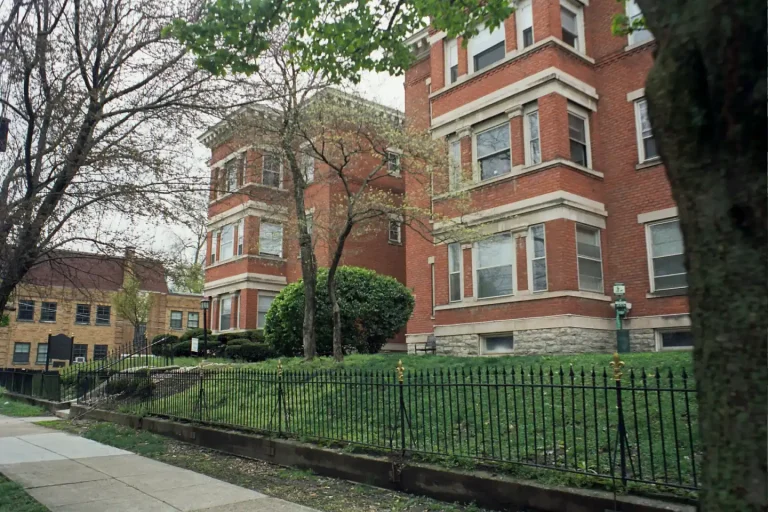 Red brick low-rise apartment building behind a black iron fence and a grassy bank on a quiet corner
