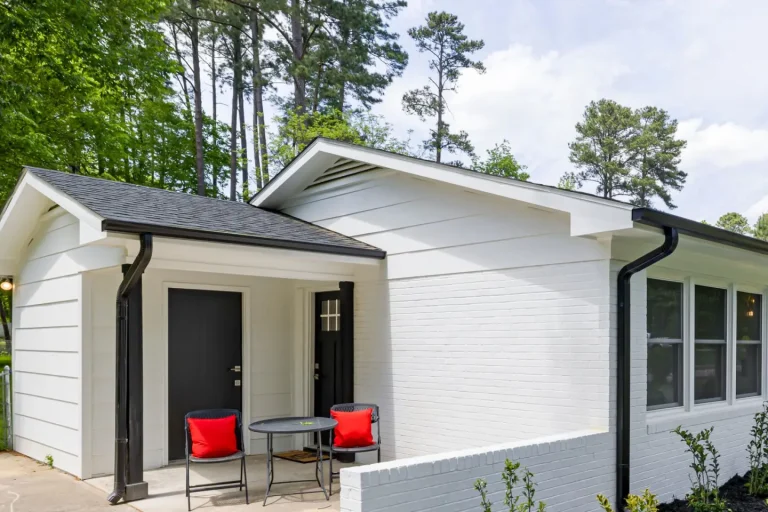 Freshly painted white brick single-storey house with two black front doors, a small patio with red chairs and new shrubs along the wall